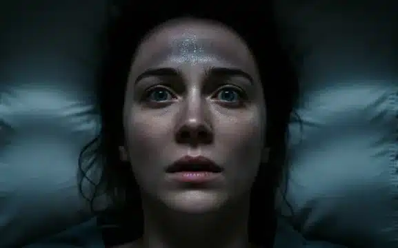 Close-up dramático de uma mulher deitada na cama à noite, com olhos arregalados e suor na testa, demonstrando sintomas de hipervigilância, ansiedade noturna e medo intenso.